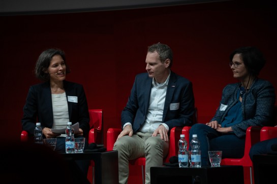 Anja Wyden Guelpa fragt die Podiumsdiskussionsteilnehmer Jochen Decker und Béatrice Vatron-Steiner eine Frage. Alle drei Diskussionsteilnehmer sitzen auf der Bühne auf roten Stühlen. Vor ihnen stehen Wasserflaschen auf schwarzen Salontischen.