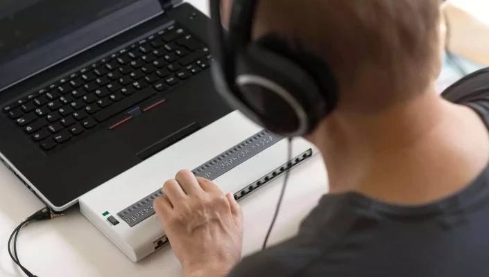 Eine Person trägt Kopfhörer und arbeitet an einem Laptop, der mit einem Braille-Display verbunden ist. Die Person bedient das Braille-Display mit der Hand, um barrierefrei auf digitale Inhalte zuzugreifen.